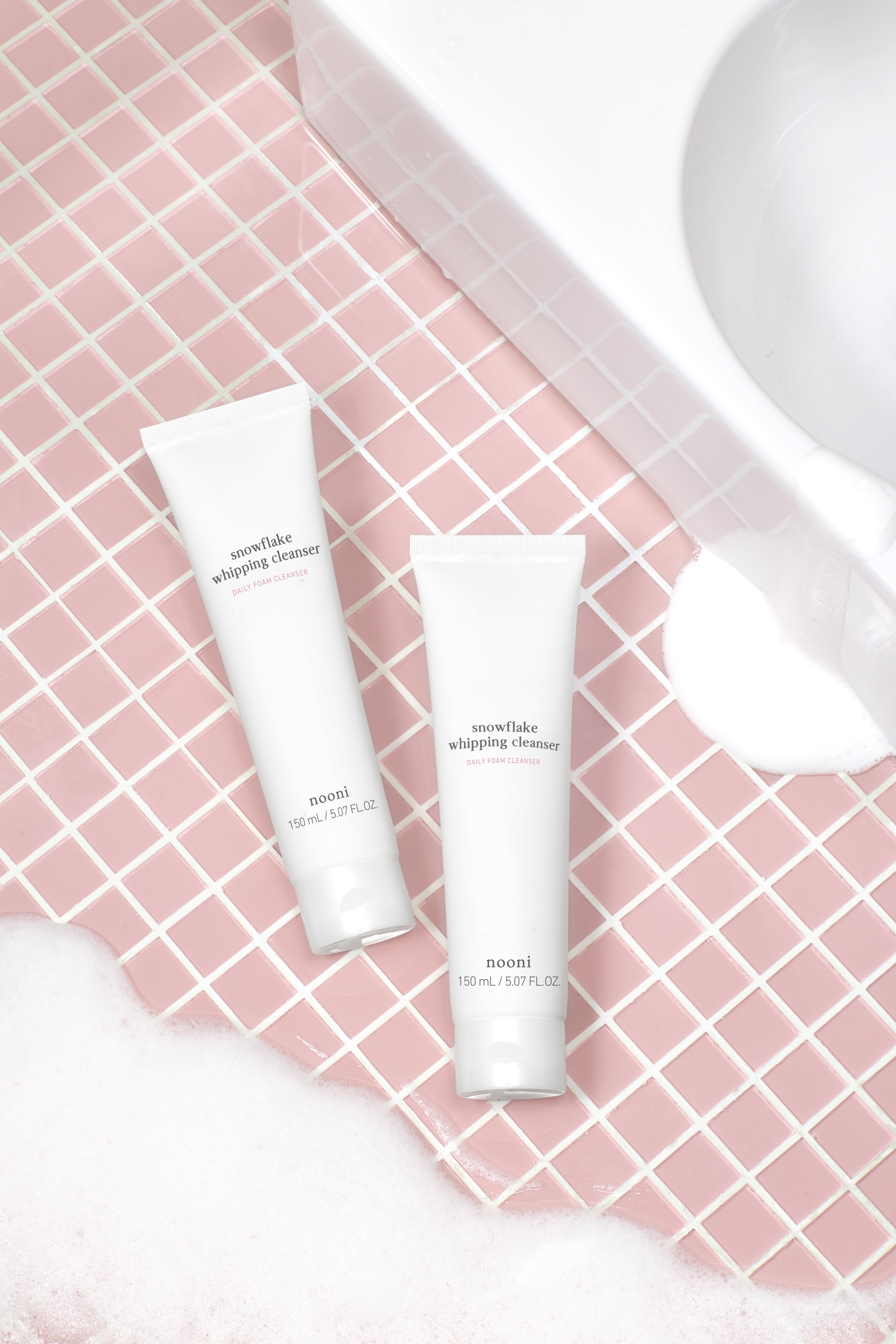 Two Nooni Snowflake Whipping Cleanser tubes in a pink tile bathroom setting with foam