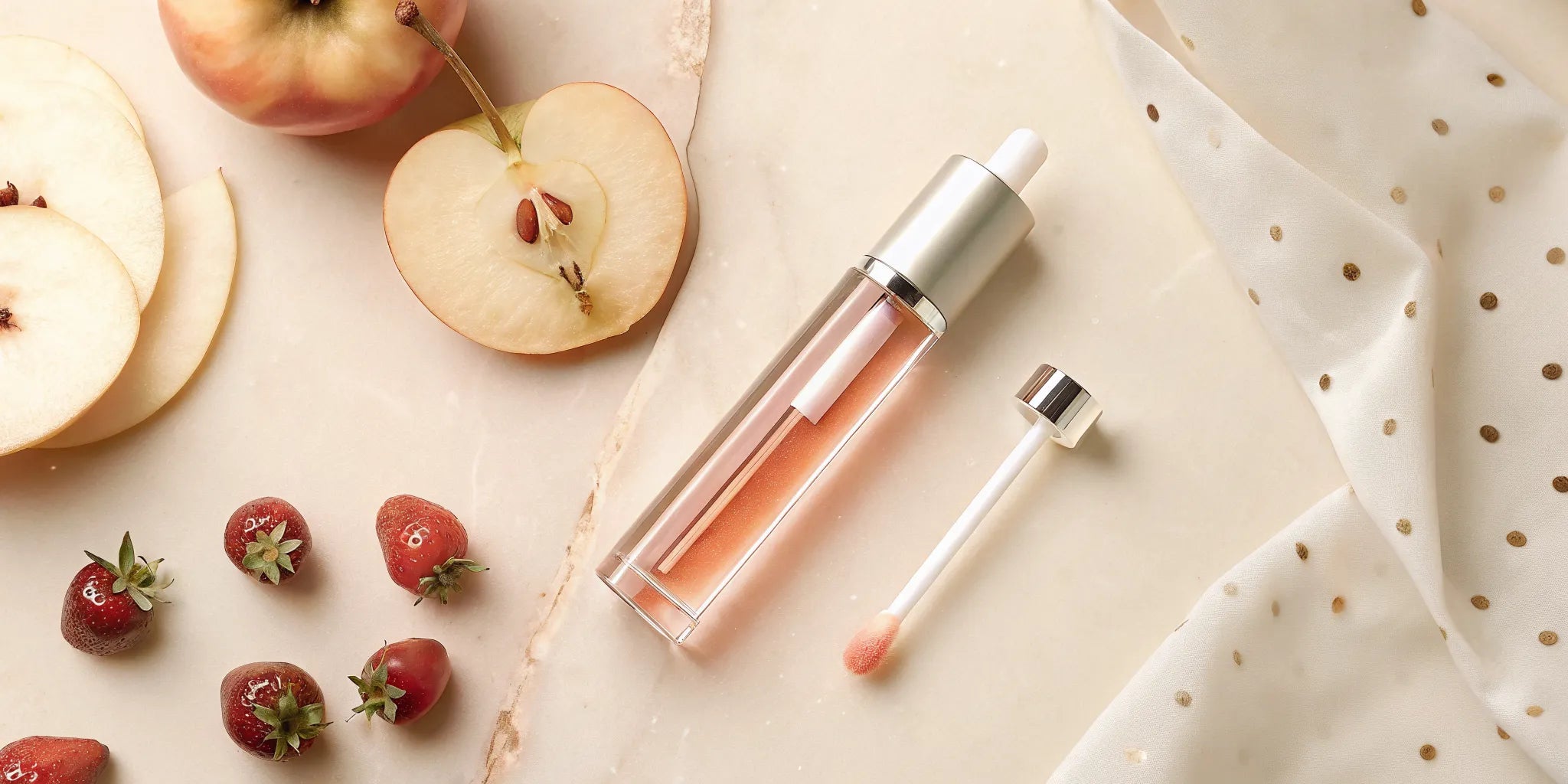 A sleek glass bottle of tinted lip oil with a doe-foot applicator, surrounded by fresh apple slices and berries on a pink marble surface